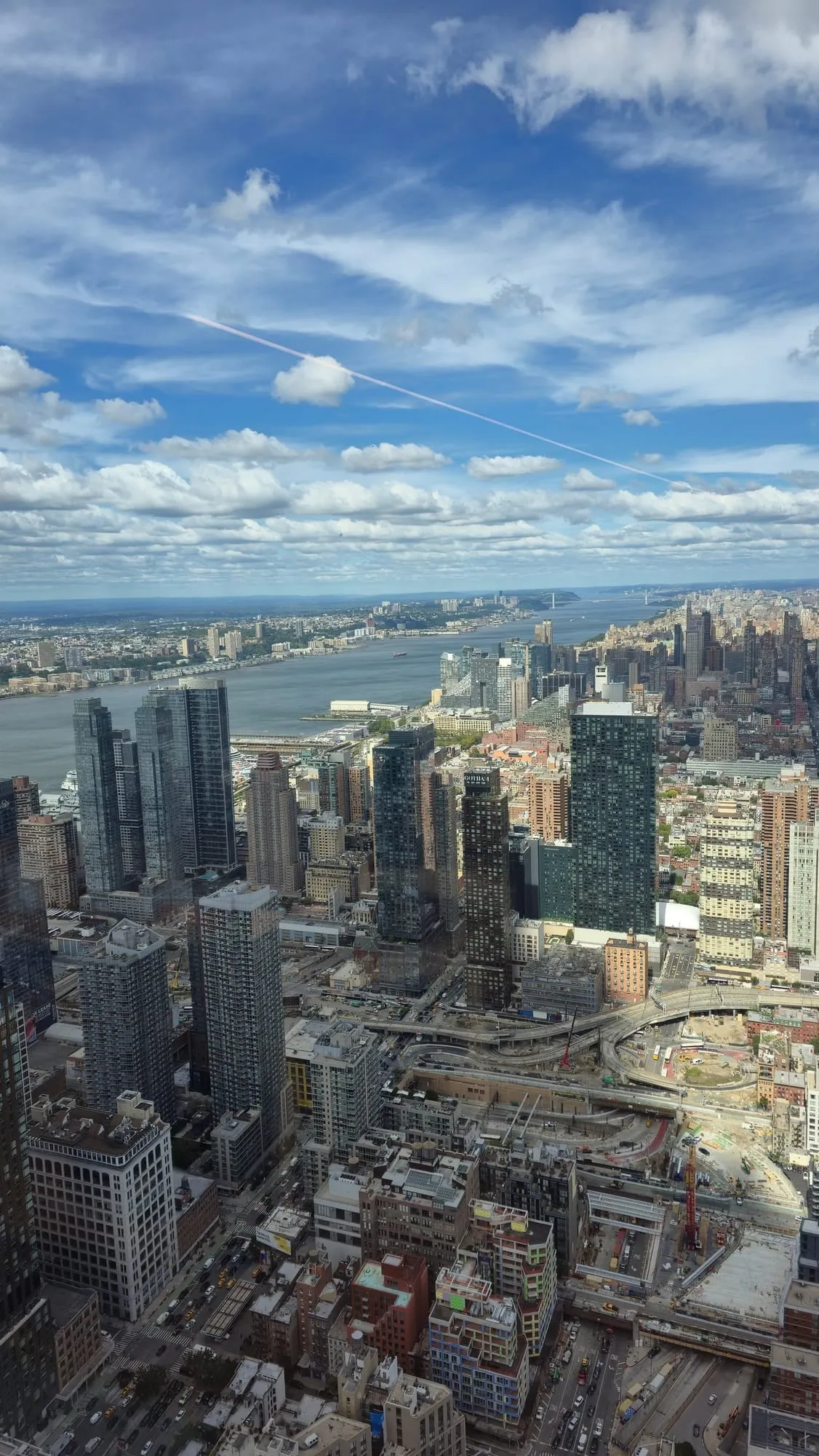 Vista desde un piso alto de Nueva York: rascacielos y calles abajo, un río al fondo y un cielo azul con nubes.