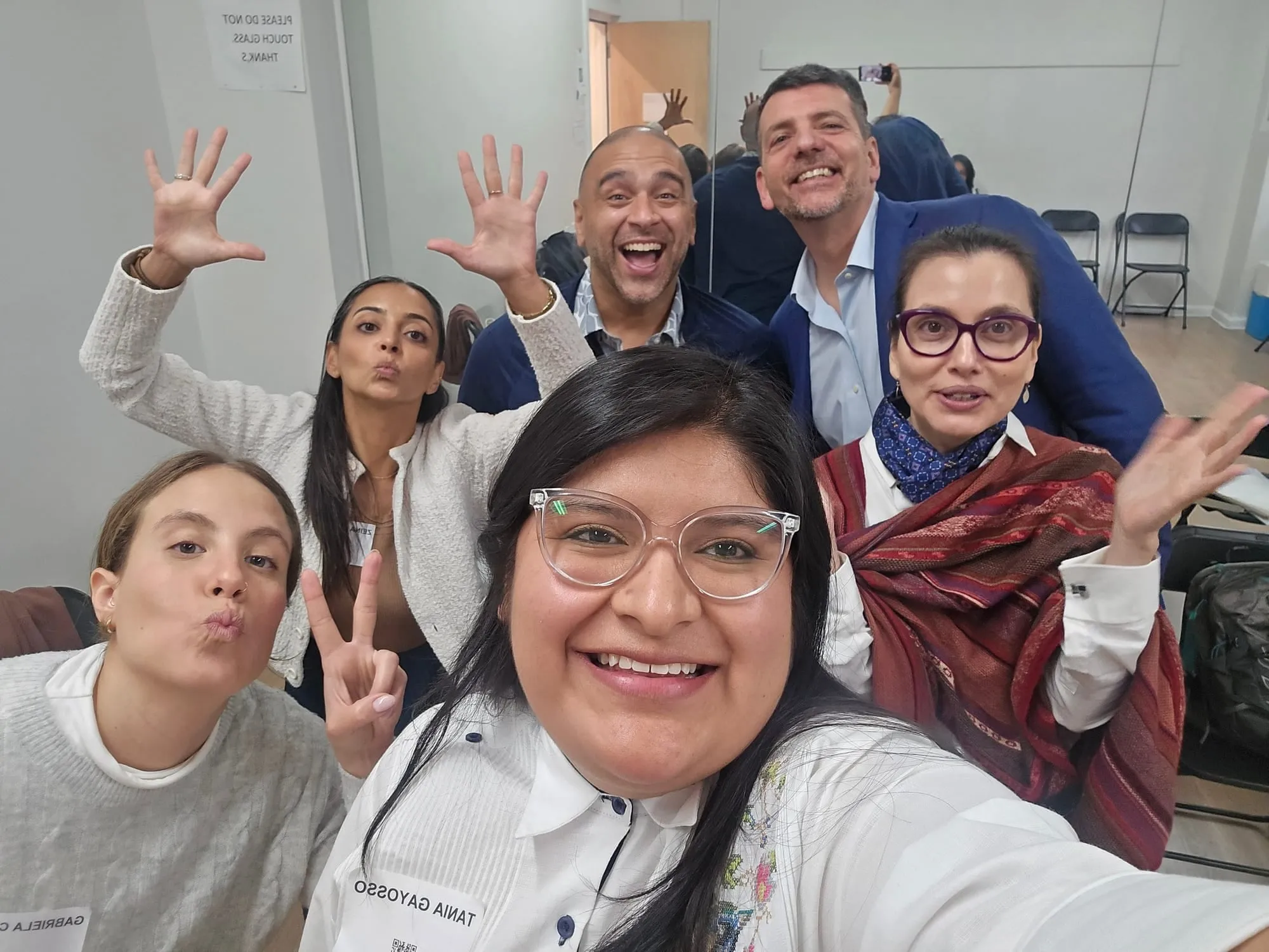 Selfi de Tania Gayosso, en primer plano y sonriendo, con cinco personas detrás que sonríen, hacen gestos con las manos o mandan besos, en un salón con sillas plegables.