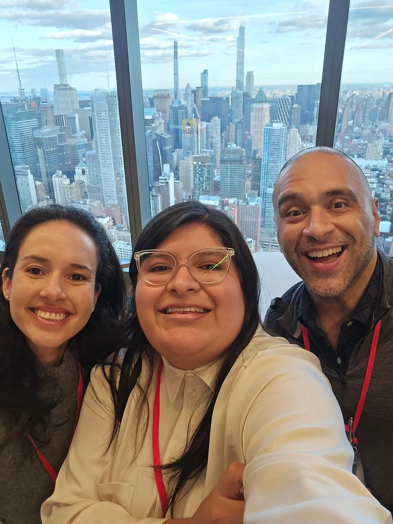 Selfi de Tania Gayosso, al centro, con una mujer a su izquierda y un hombre a su derecha; los tres sonríen frente a un ventanal con rascacielos de fondo.